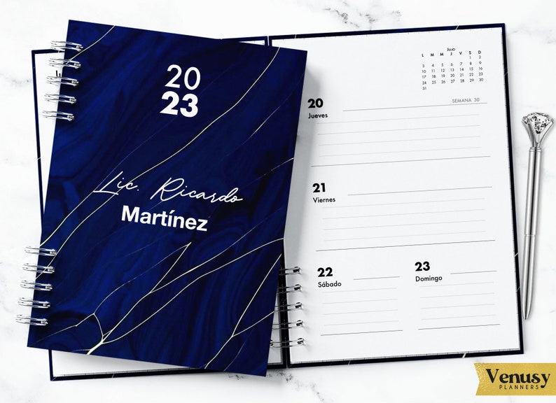 May include: A blue and gold marble-patterned planner with a white cover that says "2023" and "Lic. Ricardo Martinez". The planner is open to a weekly spread with the dates "20, 21, 22, 23" and the days of the week "Jueves, Viernes, S&aacute;bado, Domingo".