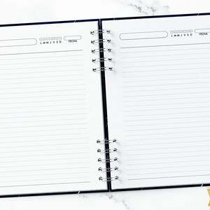 May include: An open spiral bound notebook with lined pages. The notebook has a black cover with a subtle galaxy design. The notebook is open to a blank page with a section for the day of the week and a space for the date.