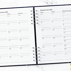 May include: A black and white planner with a silver coil binding. The planner is open to January 2023 and has a Spanish title "Planeador Enero 2023". The planner has columns for each day of the week, with spaces for notes.