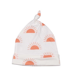 May include: White baby hat with a repeating pattern of orange suns. The hat has a knotted top and a folded brim. The suns are a solid orange color and are evenly spaced across the hat.