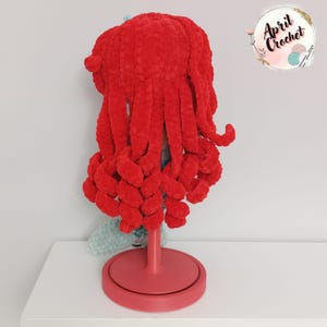 Puede incluir: Un sombrero de medusa rojo tejido a mano con largos tent&aacute;culos rizados. El sombrero se exhibe sobre un soporte rojo sobre un fondo blanco. El sombrero est&aacute; hecho de un material suave y afelpado. La imagen incluye un logotipo que dice "April Crochet".