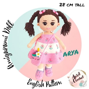 Puede incluir: Muñeca amigurumi de ganchillo llamada Arya, de 28 cm de altura, con trenzas marrones y un vestido rosa con bordados florales. La muñeca tiene un bolso a juego con un diseño de unicornio y lleva zapatos rosas. El texto "English Pattern" es visible.