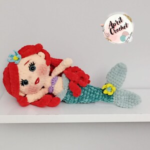 Puede incluir: Mu&ntilde;eca de sirena de ganchillo hecha a mano, con cabello rojo, ojos azules y cola turquesa. La mu&ntilde;eca tiene una flor en el pelo y un top de concha p&uacute;rpura. La mu&ntilde;eca est&aacute; tumbada en una estanter&iacute;a blanca. El logo "April Crochet" est&aacute; en la esquina superior derecha.