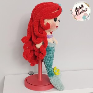 Puede incluir: Mu&ntilde;eca de ganchillo hecha a mano de una sirena con cabello rojo vibrante, una cola verde azulado y un top de concha p&uacute;rpura. La mu&ntilde;eca se exhibe en un soporte rojo. La mu&ntilde;eca tiene una corona amarilla en su cola.
