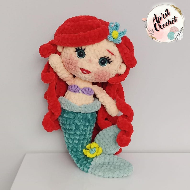 Puede incluir: Mu&ntilde;eca de sirena de ganchillo hecha a mano con cabello rojo, ojos azules y cola turquesa. La mu&ntilde;eca tiene un top de concha p&uacute;rpura y una flor azul en el cabello. Hecha de hilo suave y afelpado.