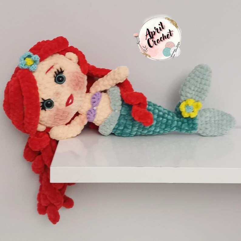 Puede incluir: Mu&ntilde;eca de sirena de ganchillo hecha a mano con cabello rojo, ojos azules y cola verde azulado. La mu&ntilde;eca lleva un top de concha p&uacute;rpura y una flor azul en el pelo. La mu&ntilde;eca est&aacute; tumbada sobre una superficie blanca.