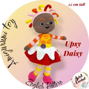 Pode incluir: Uma boneca de crochê feita à mão, Upsy Daisy, tem 22 cm de altura. A boneca apresenta uma cabeça castanha com cabelo colorido, uma parte superior laranja e amarela, uma saia vermelha com acabamento branco e sapatos rosa. O texto "Upsy Daisy" e "English Pattern" são visíveis.