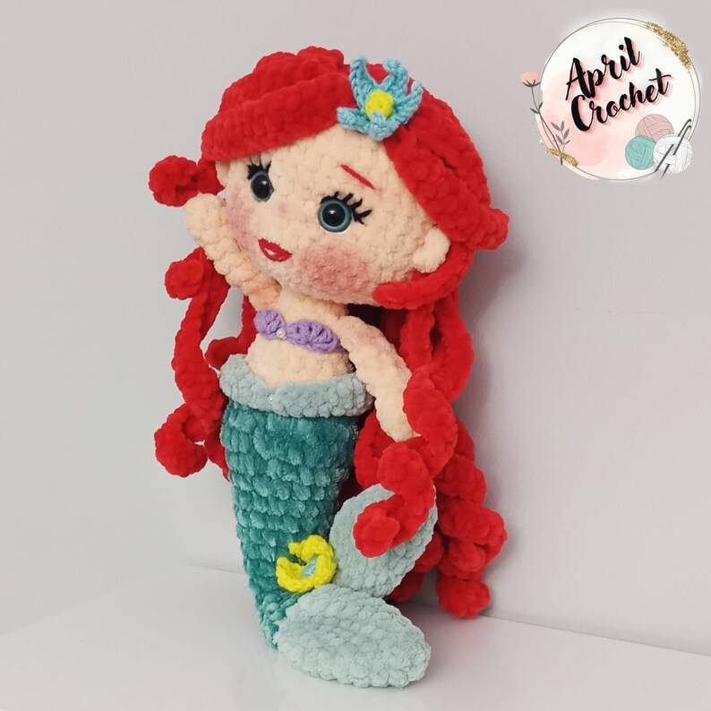 Puede incluir: Mu&ntilde;eca de sirena de ganchillo hecha a mano con cabello rojo, cola turquesa y top de concha p&uacute;rpura. La mu&ntilde;eca tiene un tono de piel claro, mejillas rosadas y una flor azul en el cabello. La mu&ntilde;eca mide aproximadamente 30 cm de alto.