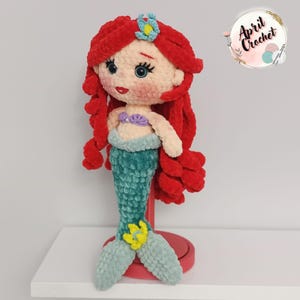 Puede incluir: Mu&ntilde;eca de sirena de ganchillo hecha a mano con cabello rojo, ojos azules y cola verde azulado. La mu&ntilde;eca tiene un top de concha p&uacute;rpura y una flor amarilla en la cola. La mu&ntilde;eca se exhibe en un soporte rojo. El texto "April Crochet" est&aacute; en la esquina superior derecha.