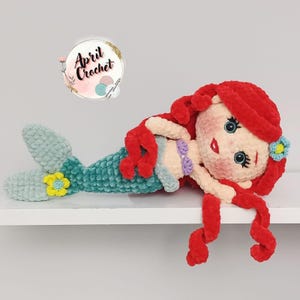 Puede incluir: Mu&ntilde;eca de sirena de ganchillo hecha a mano con cabello rojo, cola turquesa y cuerpo color crema. La sirena tiene una flor en el pelo y en la cola. La mu&ntilde;eca est&aacute; sobre una superficie blanca.