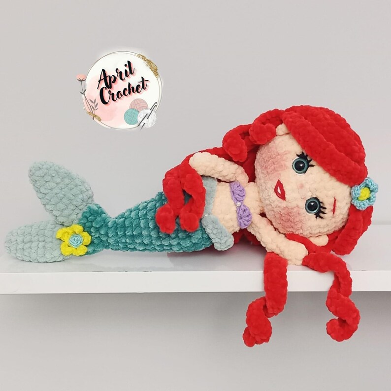 Puede incluir: Mu&ntilde;eca de sirena de ganchillo hecha a mano con cabello rojo, cola turquesa y cuerpo color crema. La sirena tiene una flor en el pelo y en la cola. La mu&ntilde;eca est&aacute; sobre una superficie blanca.