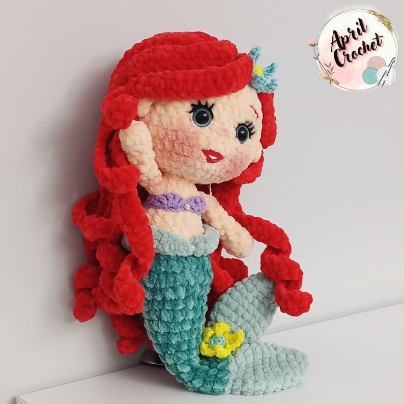 Puede incluir: Mu&ntilde;eca de sirena de ganchillo hecha a mano con cabello rojo, top morado y cola turquesa. La mu&ntilde;eca tiene una flor amarilla en la cola y lleva una flor azul en el pelo. La mu&ntilde;eca est&aacute; hecha de hilo suave y afelpado.