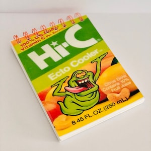 Può includere: Un piccolo quaderno verde e giallo con un design Hi-C Ecto Cooler. Il quaderno presenta un fantasma dei cartoni animati con la lingua che sporge e il testo "100% U.S.D. Vitamin C Per Serving", "Hi-C Ecto Cooler", "Citrus Drink Contains 10% Fruit Juice", e "8.45 FL. OZ. (250 mL)".
