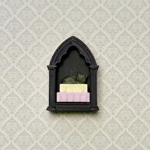 May include: A black, arched, gothic-style wall shelf featuring a miniature dark green cat figurine resting on top of two small, stacked rectangular blocks, one colored pale yellow and the other colored light pink, all displayed within the frame.