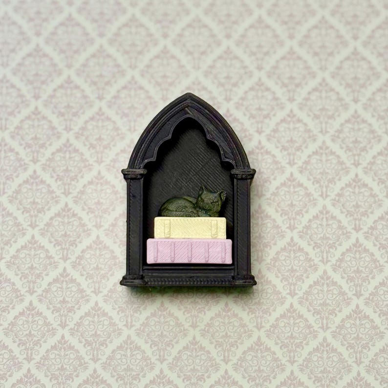 May include: A black, arched, gothic-style wall shelf featuring a miniature dark green cat figurine resting on top of two small, stacked rectangular blocks, one colored pale yellow and the other colored light pink, all displayed within the frame.