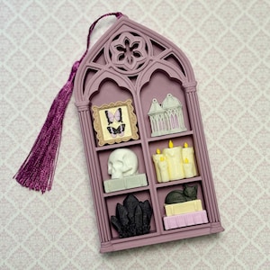 May include: A mauve gothic arch-shaped wall hanging with a purple tassel. The frame features six compartments containing miniature items: a butterfly picture, a birdcage, a white skull, yellow candles, black crystals, and a cat on books.
