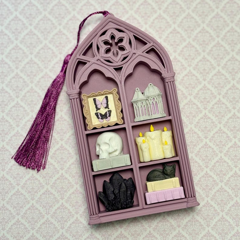 May include: A mauve gothic arch-shaped wall hanging with a purple tassel. The frame features six compartments containing miniature items: a butterfly picture, a birdcage, a white skull, yellow candles, black crystals, and a cat on books.