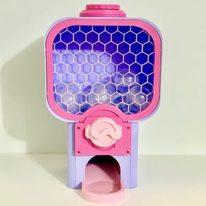 Op de afbeelding: Een kleurrijke kauwgomballenautomaat in roze, paars en blauw. De machine heeft een doorzichtige voorkant met een honingraatpatroon en een uitgiftebakje aan de onderkant. De bovenkant is effen roze.