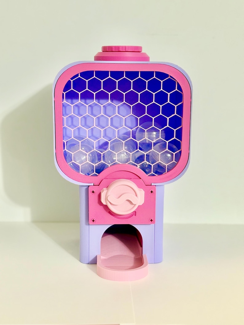 Op de afbeelding: Een kleurrijke kauwgomballenautomaat in roze, paars en blauw. De machine heeft een doorzichtige voorkant met een honingraatpatroon en een uitgiftebakje aan de onderkant. De bovenkant is effen roze.