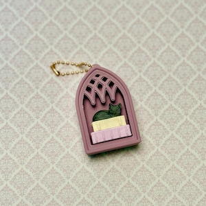 May include: A mauve keychain shaped like a gothic arched window frame. Inside the frame, a dark green cat figurine rests on top of a yellow rectangular block, which is placed above a light pink rectangular block with vertical line textures.