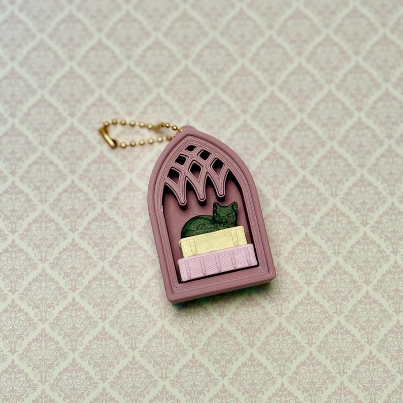 May include: A mauve keychain shaped like a gothic arched window frame. Inside the frame, a dark green cat figurine rests on top of a yellow rectangular block, which is placed above a light pink rectangular block with vertical line textures.