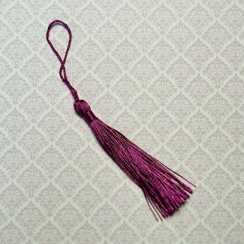 May include: A deep magenta decorative tassel featuring a tightly wrapped knot at the top and a long, flowing fringe of thin, silky threads. A matching braided cord loop extends from the top, allowing for easy attachment to various items.