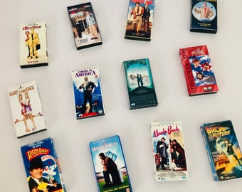 May include: A collection of VHS tapes featuring various movie titles, including "Coming to America", "Waterboy", "Uncle Buck", "Back to the Future", "Office Space", "Deeds", "Tommy Boy", "Happy Gilmore", "Princess Bride", "Box Office Hits", "Roger Rabbit", and "Don't Be a Menace to South Central While Drinking Your Juice in the Hood".