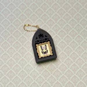 May include: A dark brown, arched, gothic-style frame keychain featuring a gold-colored rectangular inset with two small, light purple and black butterfly illustrations. The keychain includes a gold-toned ball chain for attachment.
