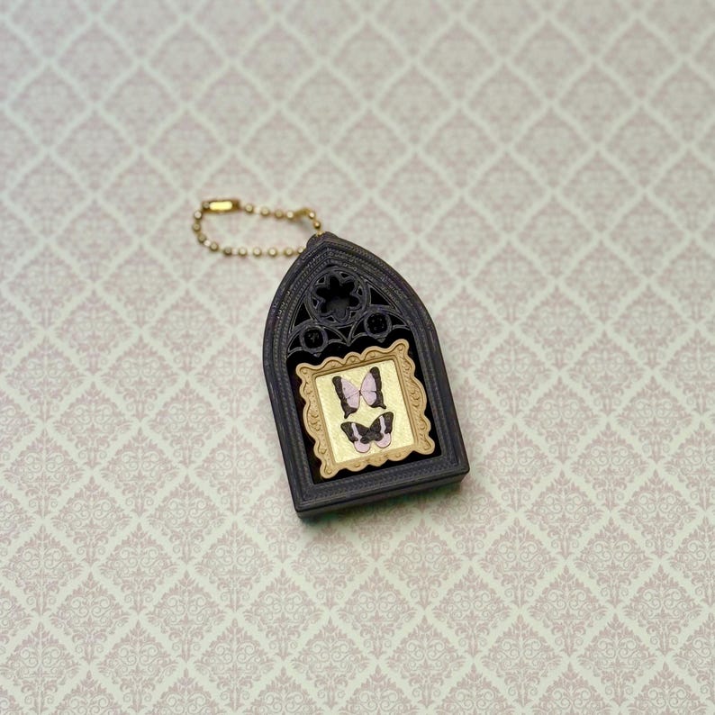 May include: A dark brown, arched, gothic-style frame keychain featuring a gold-colored rectangular inset with two small, light purple and black butterfly illustrations. The keychain includes a gold-toned ball chain for attachment.