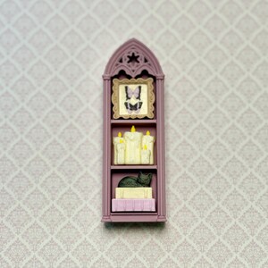 May include: A miniature mauve gothic-style arched shelf featuring three levels. The top shelf holds a framed butterfly illustration. The middle shelf displays a cluster of cream-colored pillar candles with yellow flames. The bottom shelf holds a black cat figurine resting on two stacked books.