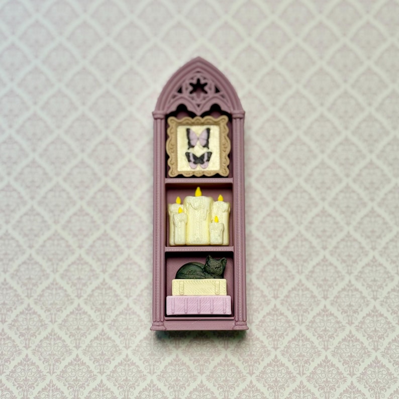 May include: A miniature mauve gothic-style arched shelf featuring three levels. The top shelf holds a framed butterfly illustration. The middle shelf displays a cluster of cream-colored pillar candles with yellow flames. The bottom shelf holds a black cat figurine resting on two stacked books.