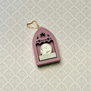 May include: A mauve-colored, arched pendant featuring a detailed, off-white skull resting on a rectangular base inside a gothic-style frame. The pendant includes a small gold-toned ball chain attachment at the top for hanging or display purposes.