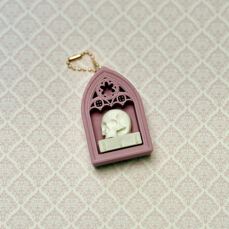 May include: A mauve-colored, arched pendant featuring a detailed, off-white skull resting on a rectangular base inside a gothic-style frame. The pendant includes a small gold-toned ball chain attachment at the top for hanging or display purposes.