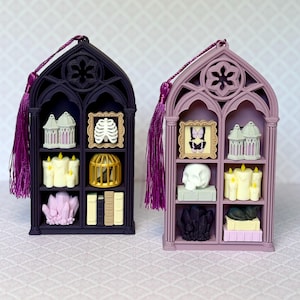 May include: Two gothic-style arched display shelves with purple tassels. The dark purple shelf holds a birdcage, a ribcage frame, candles, a gold cage, and a crystal cluster. The light purple shelf holds a butterfly frame, a birdcage, a skull, candles, and a cat.