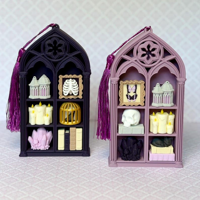 May include: Two gothic-style arched display shelves with purple tassels. The dark purple shelf holds a birdcage, a ribcage frame, candles, a gold cage, and a crystal cluster. The light purple shelf holds a butterfly frame, a birdcage, a skull, candles, and a cat.