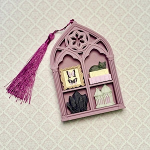 May include: A mauve, gothic-style arched wall hanging with a matching purple tassel. The frame features four compartments containing a gold-framed butterfly picture, a cat on books, a cluster of black crystals, and a small white gazebo structure.