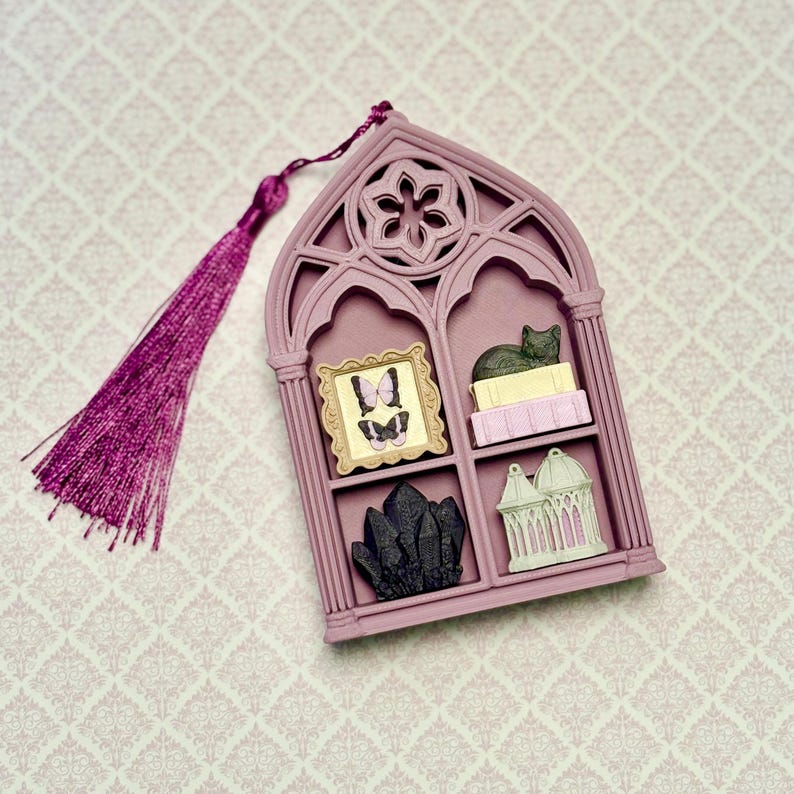 May include: A mauve, gothic-style arched wall hanging with a matching purple tassel. The frame features four compartments containing a gold-framed butterfly picture, a cat on books, a cluster of black crystals, and a small white gazebo structure.