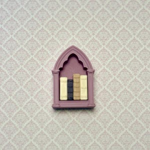 May include: A miniature mauve-colored bookshelf with a pointed gothic arch top. The shelf contains four small, upright books with textured spines in shades of cream, dark gray, tan, and off-white, arranged side by side within the frame.