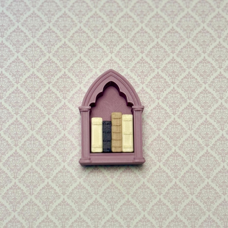 May include: A miniature mauve-colored bookshelf with a pointed gothic arch top. The shelf contains four small, upright books with textured spines in shades of cream, dark gray, tan, and off-white, arranged side by side within the frame.