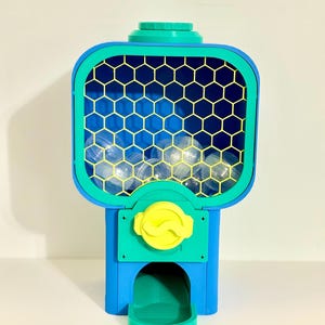 Op de afbeelding: Een kleurrijke kauwgomballenautomaat in blauw, groen en geel. De machine heeft een raam met honingraatpatroon gevuld met doorzichtige kauwgomballen. Een gele knop en een groene uitgiftebak maken het ontwerp compleet.