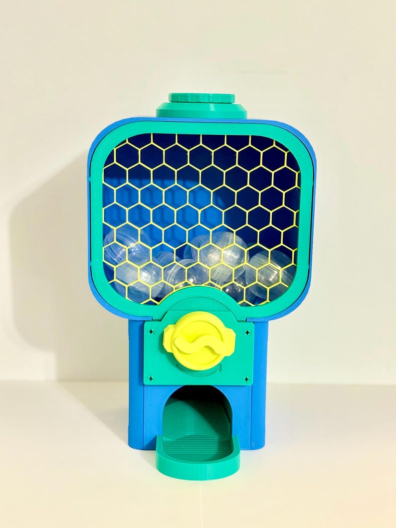 Op de afbeelding: Een kleurrijke kauwgomballenautomaat in blauw, groen en geel. De machine heeft een raam met honingraatpatroon gevuld met doorzichtige kauwgomballen. Een gele knop en een groene uitgiftebak maken het ontwerp compleet.