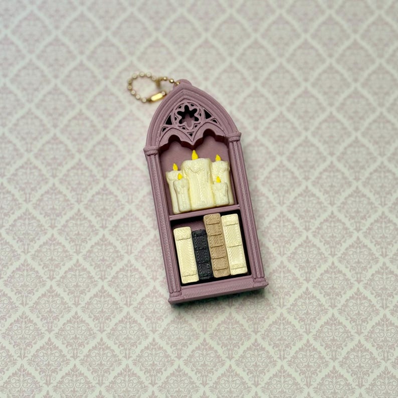 May include: A mauve-colored, arched, gothic-style keychain charm featuring a two-shelf display. The top shelf holds three cream-colored candles with yellow flames, while the bottom shelf contains four small, upright books in cream, black, and tan.