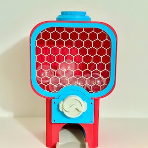 Op de afbeelding: Een rood-blauwe kauwgomballenautomaat met een doorzichtige plastic container gevuld met doorzichtige ballen. De machine heeft een blauwe bovenkant en een wit honingraatpatroon in de container. De muntsleuf is wit.