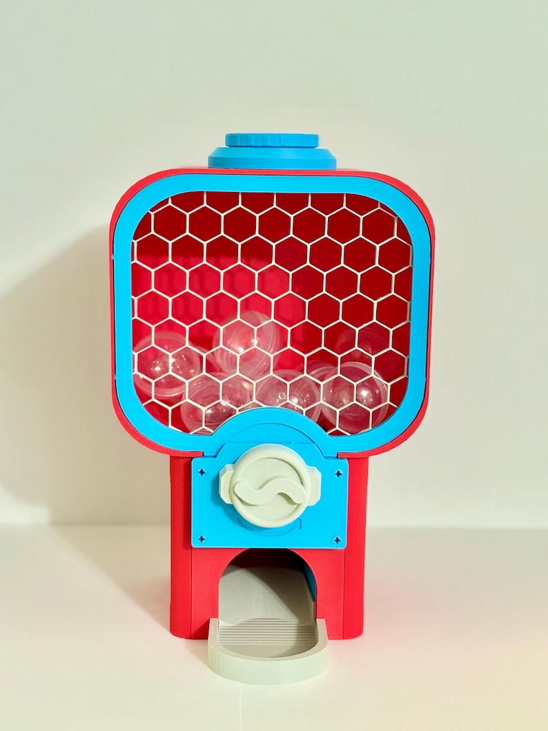 Op de afbeelding: Een rood-blauwe kauwgomballenautomaat met een doorzichtige plastic container gevuld met doorzichtige ballen. De machine heeft een blauwe bovenkant en een wit honingraatpatroon in de container. De muntsleuf is wit.