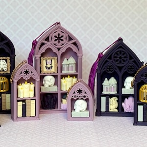 May include: A collection of miniature gothic-style arched shelves in shades of black and dusty purple, each containing small decorative items including skulls, books, candles, birdcages, and framed portraits, with some units featuring hanging tassels.