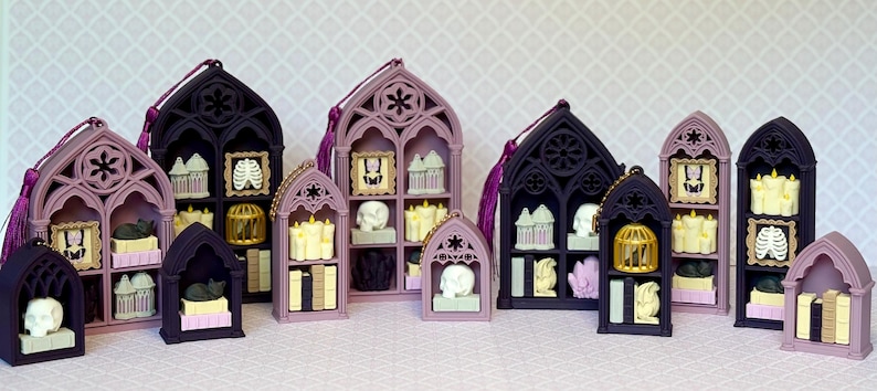 May include: A collection of miniature gothic-style arched shelves in shades of black and dusty purple, each containing small decorative items including skulls, books, candles, birdcages, and framed portraits, with some units featuring hanging tassels.