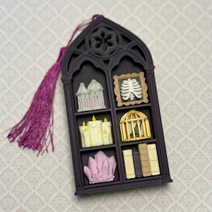 May include: A dark purple gothic window-shaped ornament with a purple tassel. The frame contains six miniature items: a white lantern, a framed white ribcage, three cream candles, a gold birdcage, pink crystals, and four small vertical books.