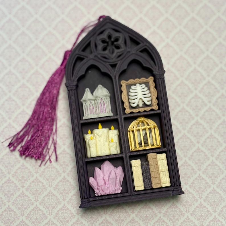 May include: A dark purple gothic window-shaped ornament with a purple tassel. The frame contains six miniature items: a white lantern, a framed white ribcage, three cream candles, a gold birdcage, pink crystals, and four small vertical books.