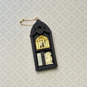 May include: A black arched pendant featuring a gold birdcage with a small bird inside on the top shelf and a white gargoyle figurine next to a small book on the bottom shelf. The pendant is attached to a gold-toned metal ball chain.