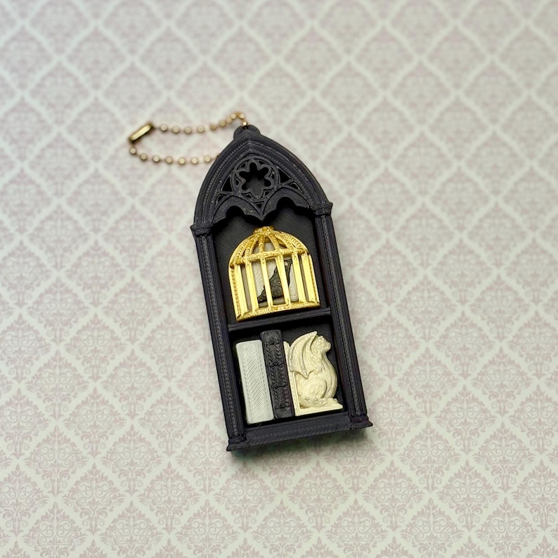 May include: A black arched pendant featuring a gold birdcage with a small bird inside on the top shelf and a white gargoyle figurine next to a small book on the bottom shelf. The pendant is attached to a gold-toned metal ball chain.